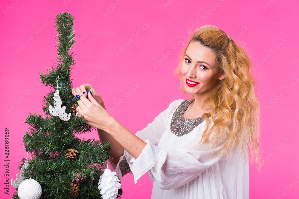 People, holidays and christmas concept - young woman hanging christmas ball on christmas tree on pink backgroung People, holidays and christmas concept - young woman hanging christmas ball on christmas tree on pink backgroung
