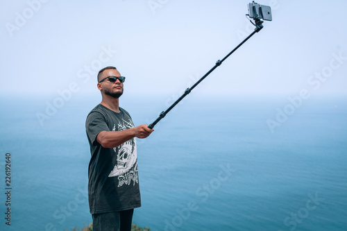 Summer photo of attractive guy in sunglasses takes a selfie on cell phone, while sitting on the bench against the sea, wearing t-shirt and shorts.