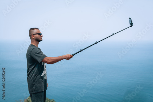 Summer photo of attractive guy in sunglasses takes a selfie on cell phone, while sitting on the bench against the sea, wearing t-shirt and shorts.