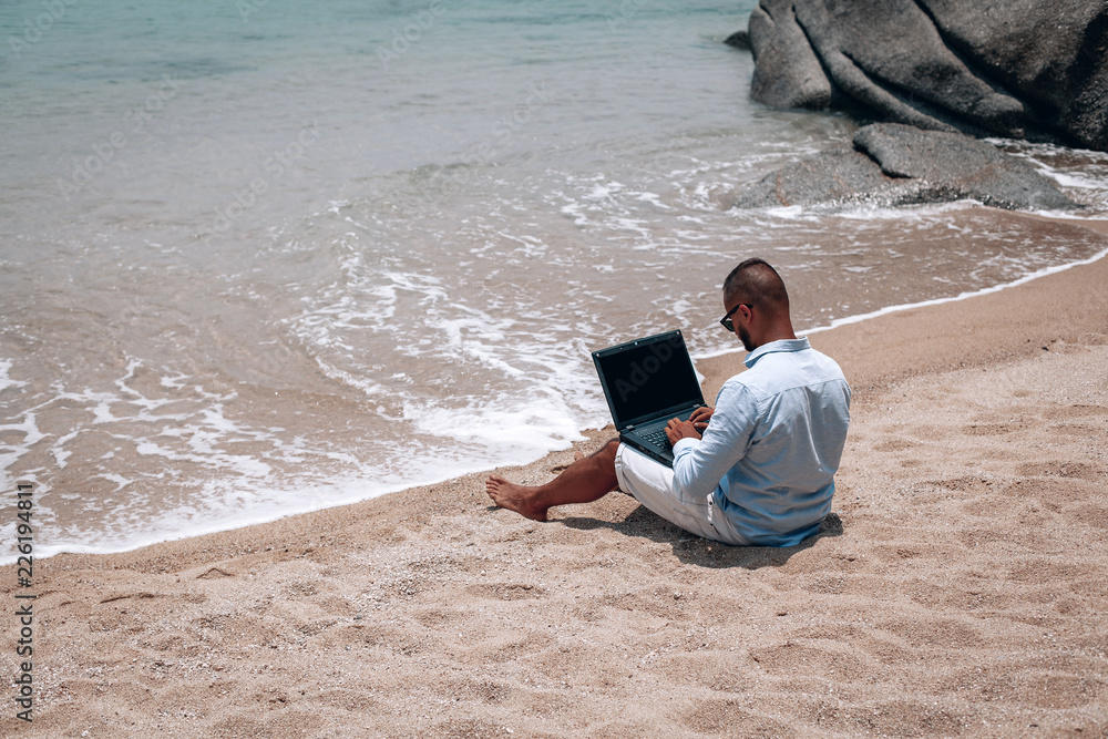 young businessman in sunglasses blue shirt and shorts working with laptop on the beach