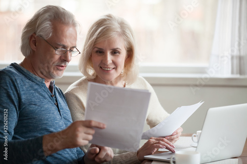 Happy old middle aged couple holding reading good news in document, smiling senior mature family excited by mail letter, checking paying domestic bills online on laptop, discussing budget planning