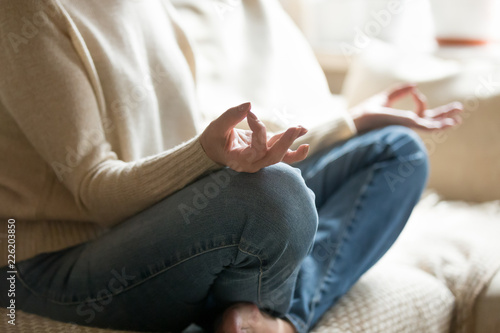 Calm senior woman sitting on sofa holding hands in mudra practicing home yoga in lotus pose for relaxation, old mature middle aged female enjoying meditating alone for peaceful mind, close up view