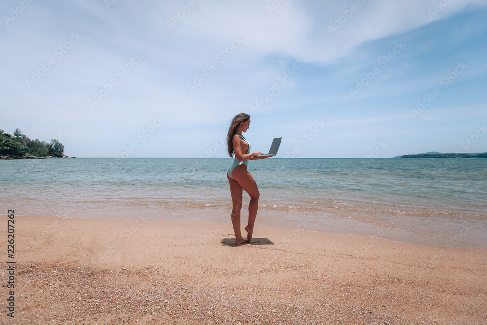 Young american woman in blue swimsuit working with laptop on empty beach. Concept of resting on morning sea and summer vacations, modern technology. Young american woman in blue swimsuit working with laptop on empty beach. Concept of resting on morning sea and summer vacations, modern technology.