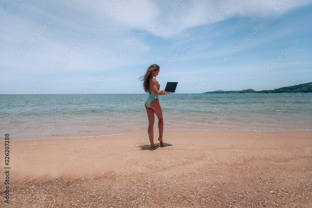 Young american woman in blue swimsuit working with laptop on empty beach. Concept of resting on morning sea and summer vacations, modern technology. Young american woman in blue swimsuit working with laptop on empty beach. Concept of resting on morning sea and summer vacations, modern technology.