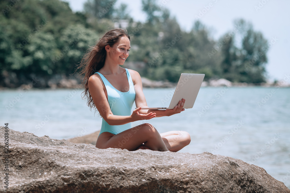 Young beatiful  woman working with laptop on empty beach and sitting on stone, showing thumbs up. Concept of resting on morning sea and summer vacations, modern technology.