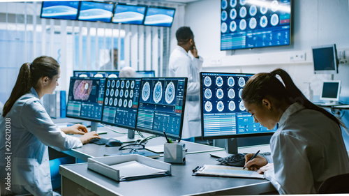 Team of Professional Scientists Work in the Brain Research Laboratory. Neurologists / Neuroscientists Surrounded by Monitors Showing CT, MRI Scans Having Discussions and Working on Personal Computers.