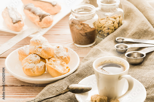 Home cooked profiteroles and eclairs with a cup of black coffee on a wooden table.