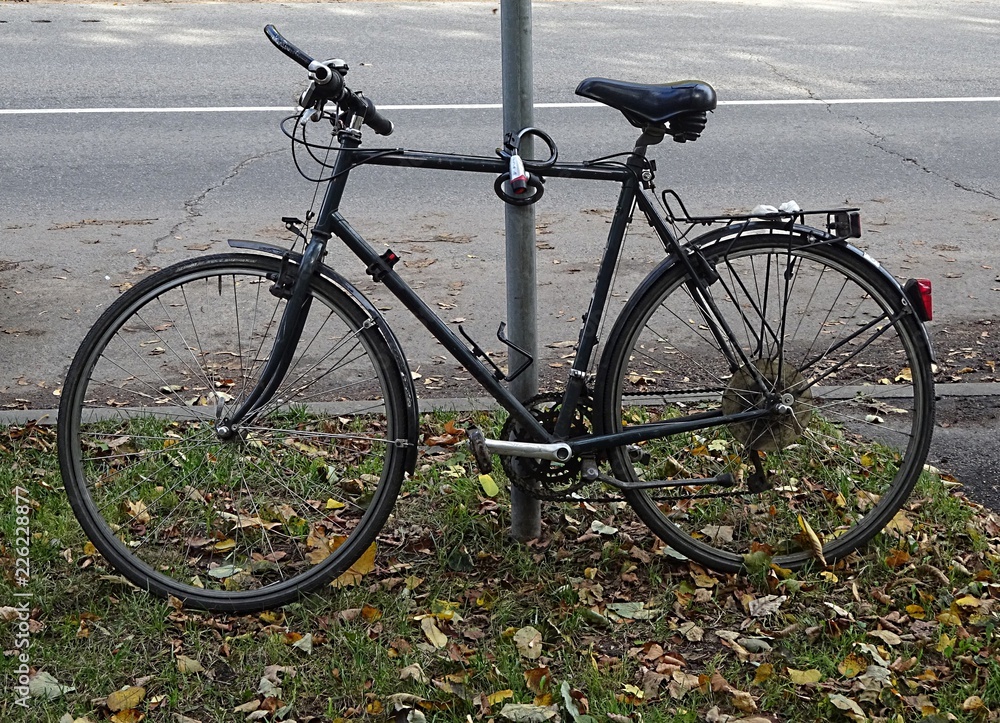 Fototapeta premium Black Mountain City Multi Speed Road Bike Cycle Bicycle With Anti Theft Lock Parked On The Green Grass With Leafs Behind Asphalt 