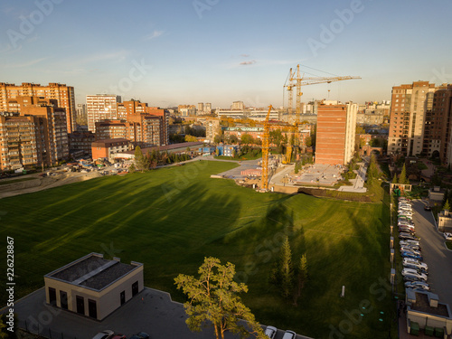 Aerial view of landscape with an elite building in the center of the city with skyscraper and three large yellow cranes with a large lawn of green grass in the fall during sunset on a clear day photo