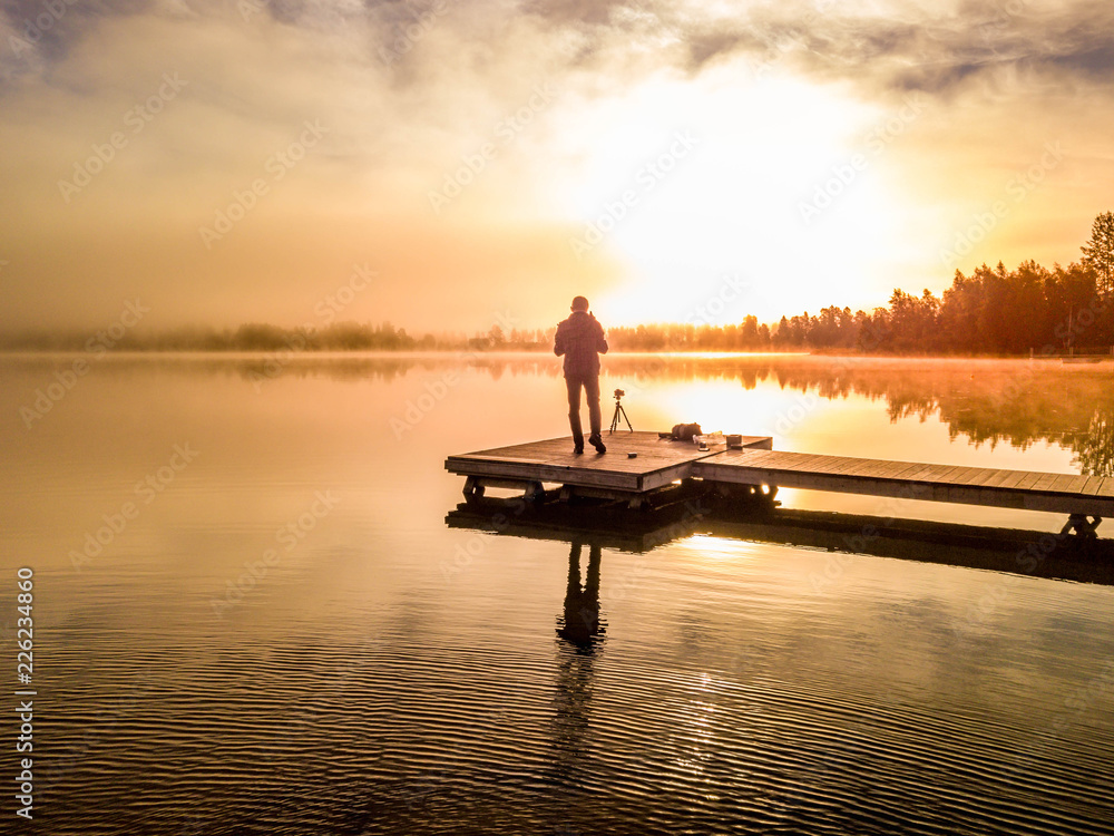 operator of drone drives it to film and take photos of films foggy bright sunrise over lake and trees, standing with his back to us, West Bothnia province, north of Sweden