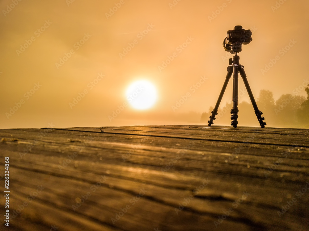 tripod photo camera on wooden bridge films bright sunrise through over lake and trees very thick golden fog, West Bothnia province, north of Sweden tripod photo camera on wooden bridge films bright sunrise through over lake and trees very thick golden fog, West Bothnia province, north of Sweden