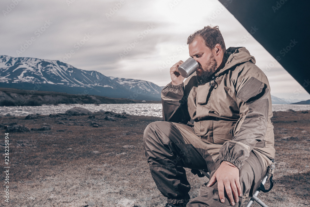 Young man drinking tea or coffee in mountains. Mountain camping concept. Traveler man with beard sit near the mountain lake . Man traveler hands holding cup of tea. the view from the tent