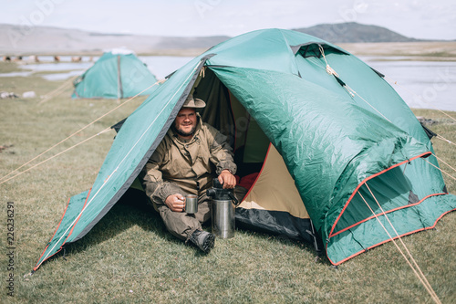 Young man in hat, drinking tea or coffee in mountains. Mountain camping concept. Traveler man with beard sit near the tent neat the mountain lake . Man traveler hands holding cup of tea.