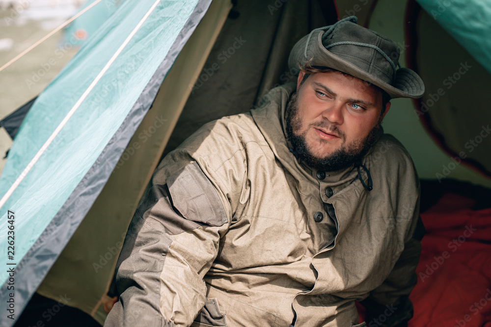 Fototapeta premium Young man in hat, drinking tea or coffee in mountains. Mountain camping concept. Traveler man with beard sit near the tent neat the mountain lake . Man traveler hands holding cup of tea.