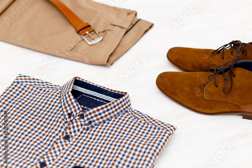 Beige pants, plaid shirt, brown suede shoes and leather belt. Overhead view of men's casual outfit on white wooden background.