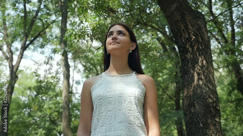 Down up view of a slender young woman with long loose hair looking up in a green forestry full of shining rays on a sunny day in summer Down up view of a slender young woman with long loose hair looking up in a green forestry full of shining rays on a sunny day in summer