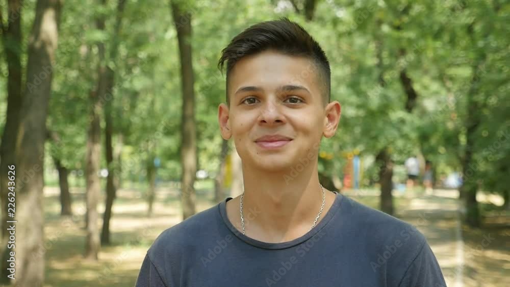 Portrait of a young brunet man with a short haircut standing and smiling cheery in a green park ull of leafy trees on a sunny day Portrait of a young brunet man with a short haircut standing and smiling cheery in a green park ull of leafy trees on a sunny day