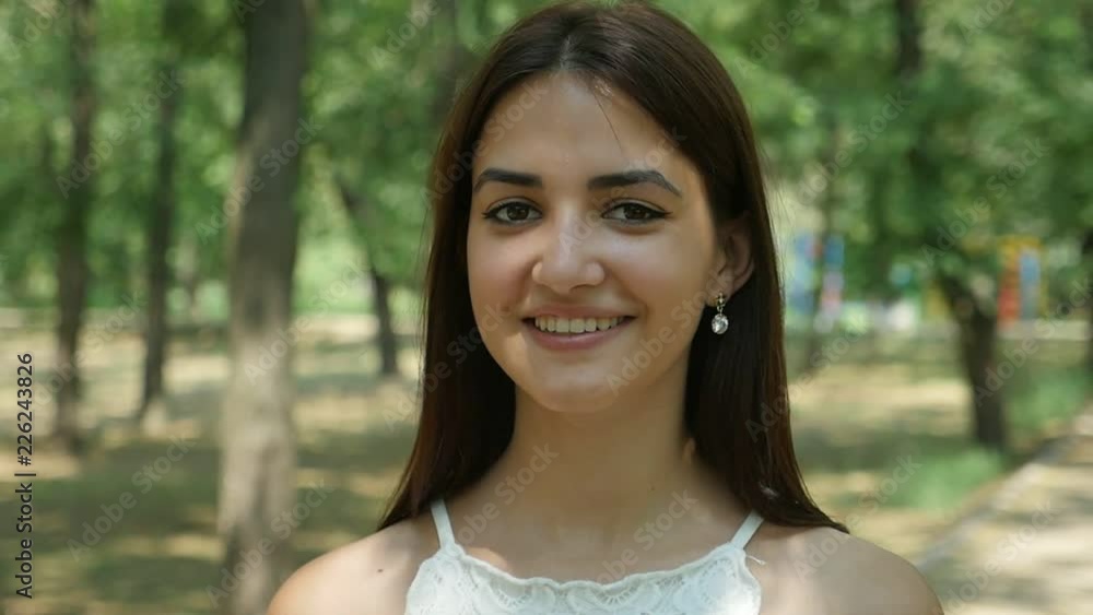 Portrait of a cheery young woman with long loose hair standing and smiling happily in a leafy park with full of sun rays on a sunny day in summer Portrait of a cheery young woman with long loose hair standing and smiling happily in a leafy park with full of sun rays on a sunny day in summer