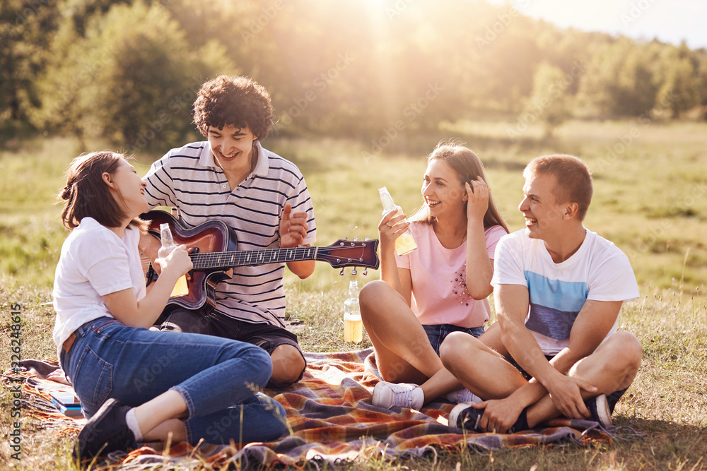 Summer, vacation, music and recreation time concept. Cheerful four friends  or classmates have picnic outdoor, sing songs to guitar, drink energetic  beverages, enjoys sunny warm day, pose on field Stock-Foto | Adobe