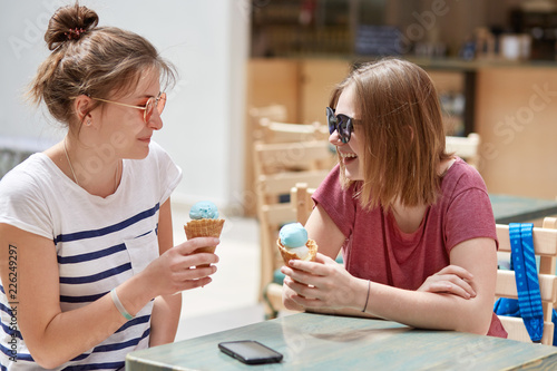 People, lifestyle and rest concept. Joyful hipsters have spare tine, eat delicious ice cream in cafe during summer, wear sunglasses, tell each other anecdotes, being in good mood, have nice relations