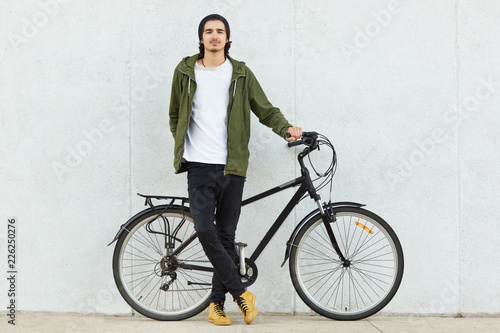 Isolated shot of stylish hipster male teenager dressed in stylish outfit and sneakers, stands near sport bicycle, enjoys recreation time alone, advertises his bike, poses over white concrete wall.