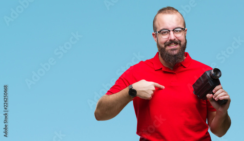 Young caucasian hipster man filmming using retro video camera over isolated background with surprise face pointing finger to himself