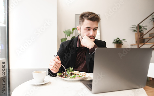 Portrait of a concentrated man sitting at a table in a cafe with a plate of salad and a cup of coffee and uses a laptop. Busy man works on a laptop in a restaurant at lunch time.