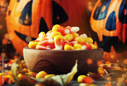 Halloween festive composition with sweet corn in bowl and smiling pumpkins guards, lantern, straw and fallen leaves on dark wooden background, rustic style, selective focus