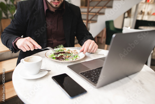 Business man eating breakfast in a restaurant. Plate of salad  a laptop  a cup of coffee  a smartphone on the table in the restaurant. Man eats a plate salad with a fork and a knife.