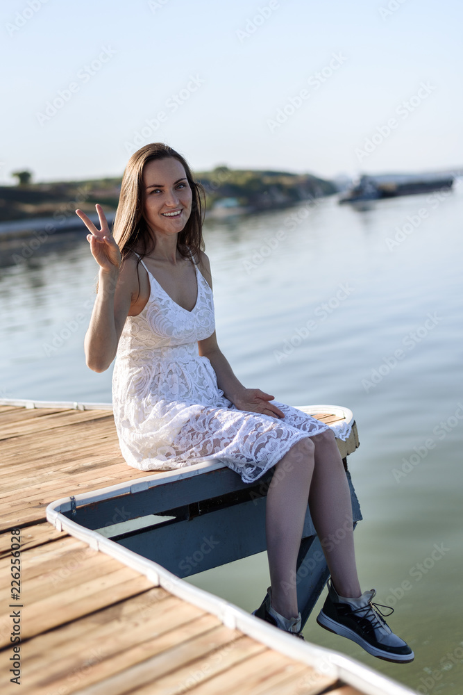 Young smiling brunette sitting on a pier in a white dress shows a hand sign of peace