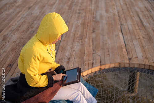 A woman in a yellow jacket sitting on a wooden pier, uses a tablet