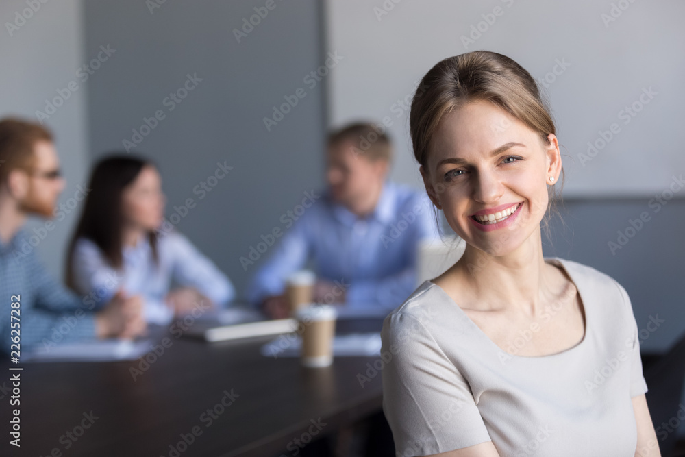 Smiling young attractive professional businesswoman looking at camera in office with colleagues at background, happy successful female business leader, coach or employee executive headshot portrait Smiling young attractive professional businesswoman looking at camera in office with colleagues at background, happy successful female business leader, coach or employee executive headshot portrait