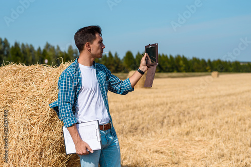 A young agronomist in a white t-shirt and a blue shirt with a tablet and a notebook stands at a haystack, taking pictures of the field