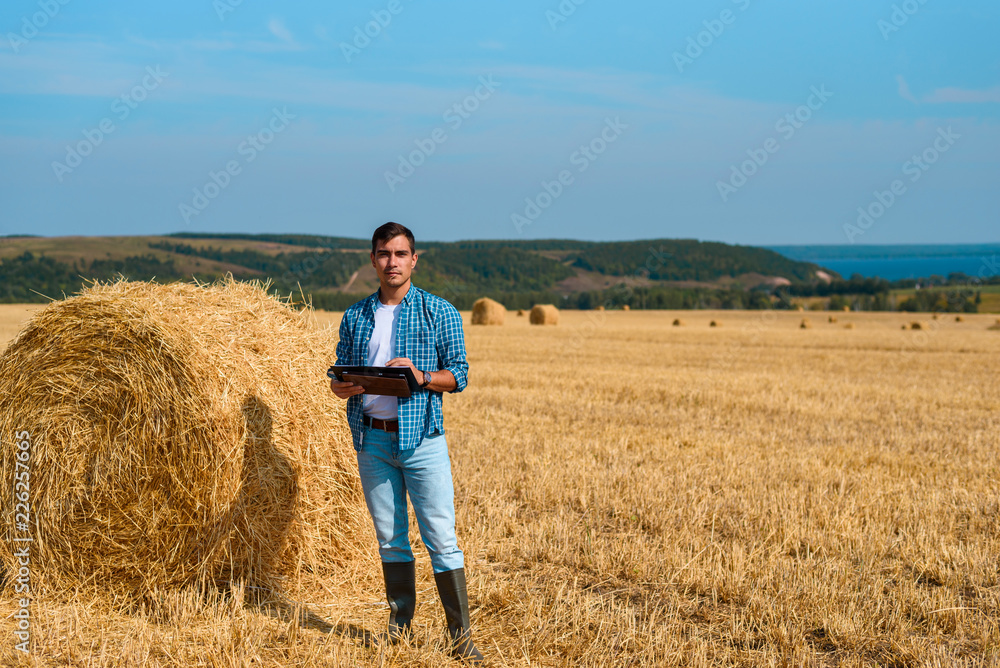 Frontal portrait of man botanist farmer with a tablet in blue jeans and a shirt and a white t-shirt in a field with haystacks Frontal portrait of man botanist farmer with a tablet in blue jeans and a shirt and a white t-shirt in a field with haystacks