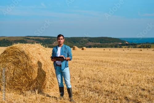 Frontal portrait of man botanist farmer with a tablet in blue jeans and a shirt and a white t-shirt in a field with haystacks © Руслан Галиуллин