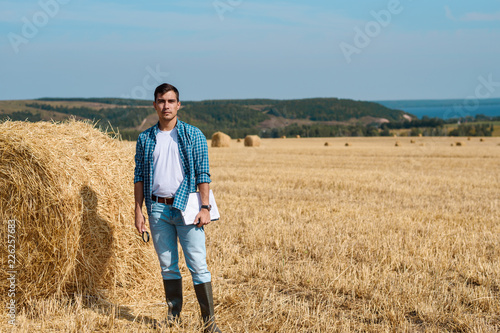 Young farmer in jeans, white t-shirt, blue shirt, rubber boots in the field with haystacks with tablet and notebook, copy space, agricultural industry, rural business concept © Руслан Галиуллин