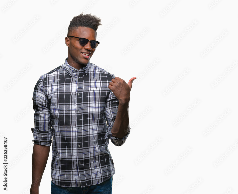 Young african american man wearing sunglasses over isolated background smiling with happy face looking and pointing to the side with thumb up. Young african american man wearing sunglasses over isolated background smiling with happy face looking and pointing to the side with thumb up.