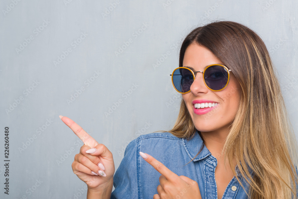 Young adult woman over grunge grey wall wearing retro sunglasses smiling and looking at the camera pointing with two hands and fingers to the side. Young adult woman over grunge grey wall wearing retro sunglasses smiling and looking at the camera pointing with two hands and fingers to the side.