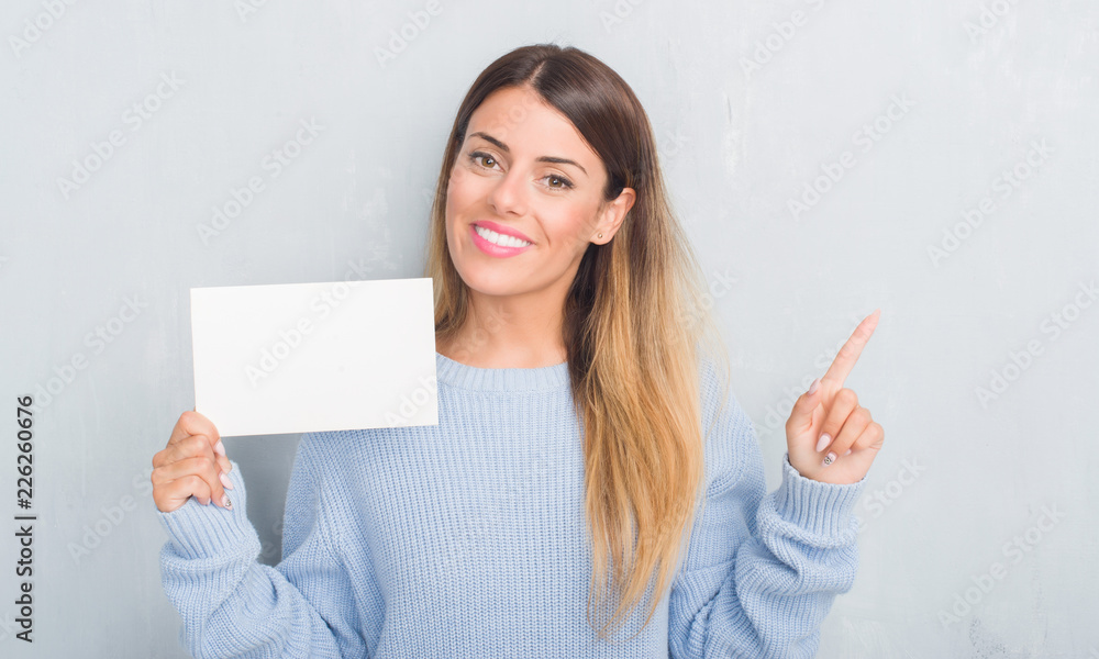 Young adult woman over grey grunge wall holding blank card very happy pointing with hand and finger to the side Young adult woman over grey grunge wall holding blank card very happy pointing with hand and finger to the side