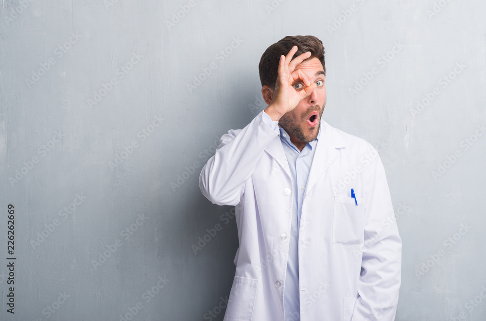Handsome young professional man over grey grunge wall wearing white coat doing ok gesture shocked with surprised face, eye looking through fingers. Unbelieving expression.