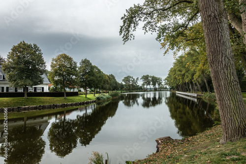 Sluis, Zeeland, Netherlands - September 22, 2018: Gray sky and green bordering trees reflected in quiet water of dead ending canal Bruges-Sluis in Sluis. White house. © Klodien
