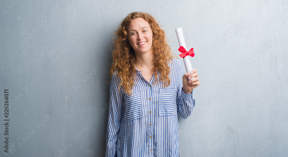 Young redhead business woman over grey grunge wall holding diploma with a happy face standing and smiling with a confident smile showing teeth Young redhead business woman over grey grunge wall holding diploma with a happy face standing and smiling with a confident smile showing teeth