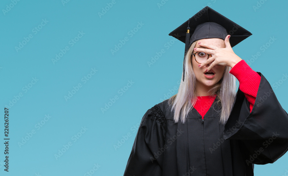 Young blonde woman wearing graduate uniform over isolated background peeking in shock covering face and eyes with hand, looking through fingers with embarrassed expression. Young blonde woman wearing graduate uniform over isolated background peeking in shock covering face and eyes with hand, looking through fingers with embarrassed expression.