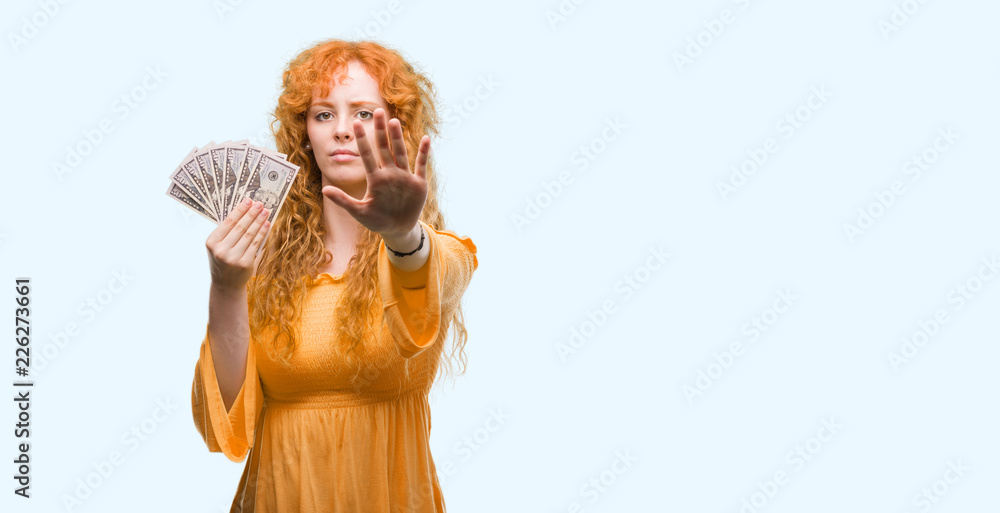 Young redhead woman holding dollars with open hand doing stop sign with serious and confident expression, defense gesture