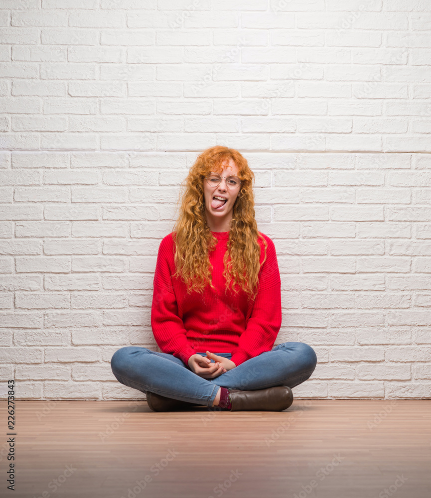 Young redhead woman sitting over brick wall sticking tongue out happy with funny expression. Emotion concept. Young redhead woman sitting over brick wall sticking tongue out happy with funny expression. Emotion concept.