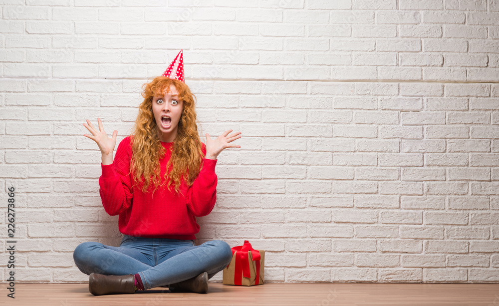 Young redhead woman sitting over brick wall wearing birthday hat very happy and excited, winner expression celebrating victory screaming with big smile and raised hands Young redhead woman sitting over brick wall wearing birthday hat very happy and excited, winner expression celebrating victory screaming with big smile and raised hands