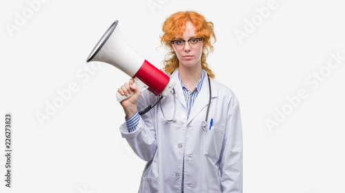 Young redhead doctor woman holding megaphone with a confident expression on smart face thinking serious
