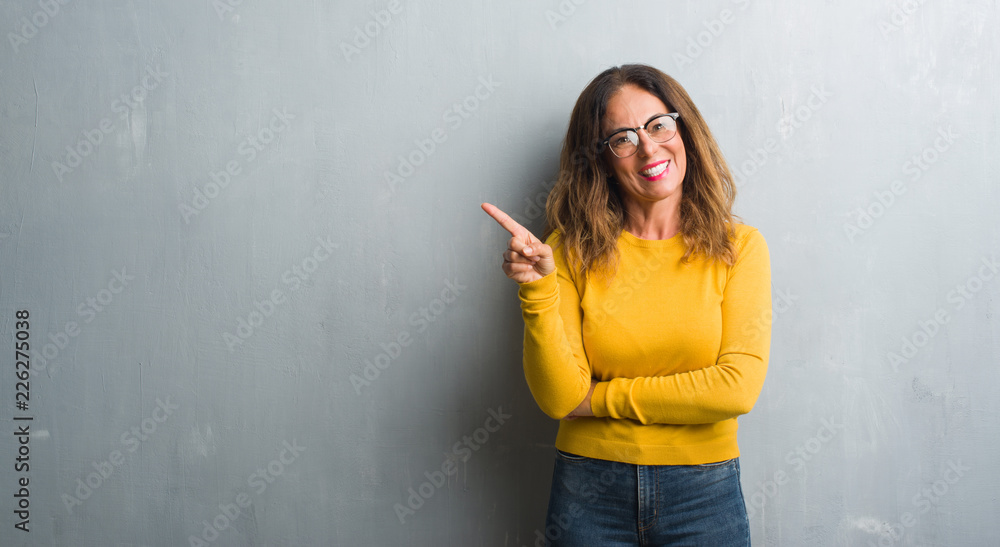 Middle age hispanic woman over grey wall wearing glasses with a big smile on face, pointing with hand and finger to the side looking at the camera. Middle age hispanic woman over grey wall wearing glasses with a big smile on face, pointing with hand and finger to the side looking at the camera.
