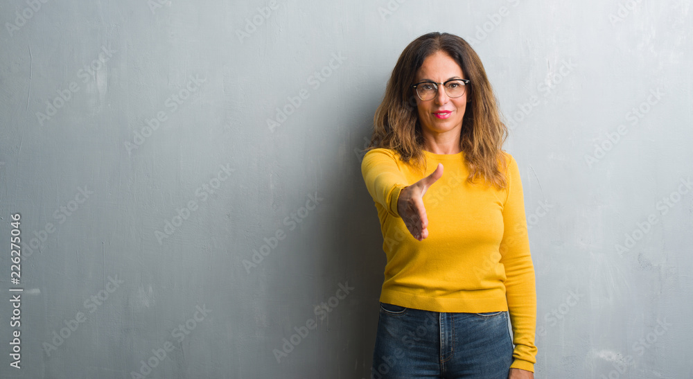Middle age hispanic woman over grey wall wearing glasses smiling friendly offering handshake as greeting and welcoming. Successful business. Middle age hispanic woman over grey wall wearing glasses smiling friendly offering handshake as greeting and welcoming. Successful business.