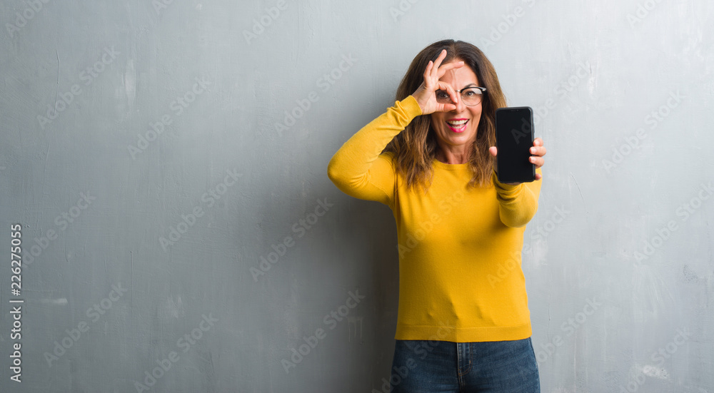 Middle age hispanic woman showing smarphone screen with happy face smiling doing ok sign with hand on eye looking through fingers